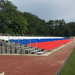 Масштабная сборно-разборная трибуна: многоярусная трибуна с сине-красными сиденьями, вид сбоку на стадионе, примыкание к беговой дорожке, металлический каркас и лестницы.