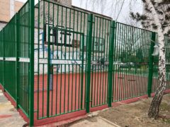 Крупный план секции металлического ограждения спортивной площадки, видны вертикальные прутья, зелёная порошковая окраска и распашные ворота