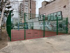 Установленное металлическое ограждение спортивной площадки зелёного цвета, видны ворота и примыкание к кирпичной стене здания