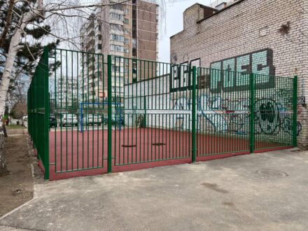 Установленное металлическое ограждение спортивной площадки зелёного цвета, видны ворота и примыкание к кирпичной стене здания