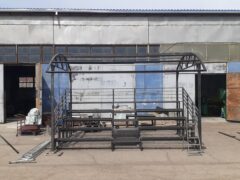 Металлический каркас трибуны с навесом, вид спереди, конструкция стоит на площадке возле промышленного здания, видны ступени и опорные стойки
